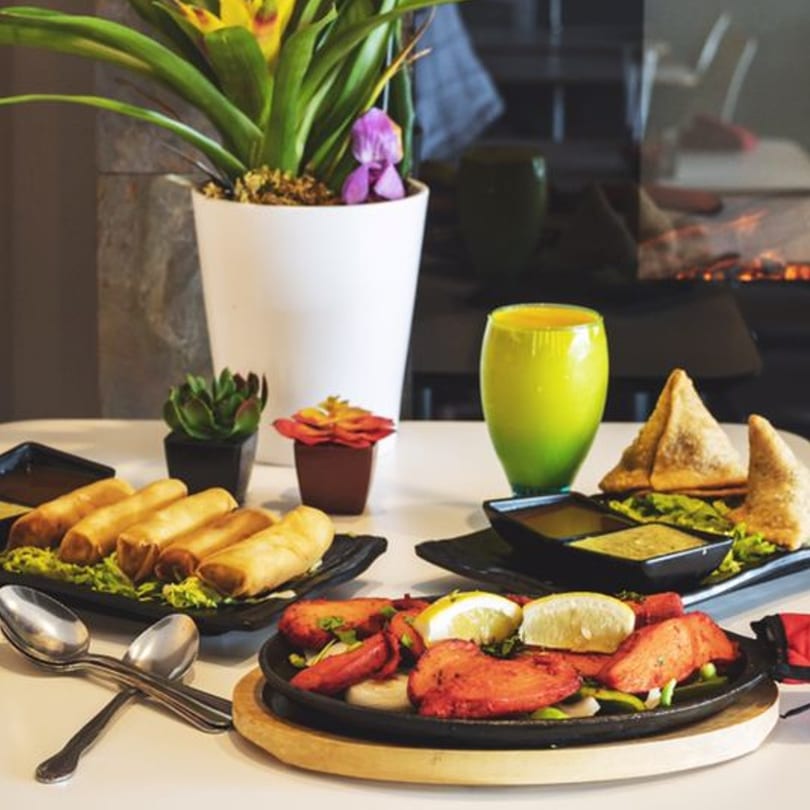 Indian appetizers including samosas, spring rolls, and tandoori chicken on a table with plants.