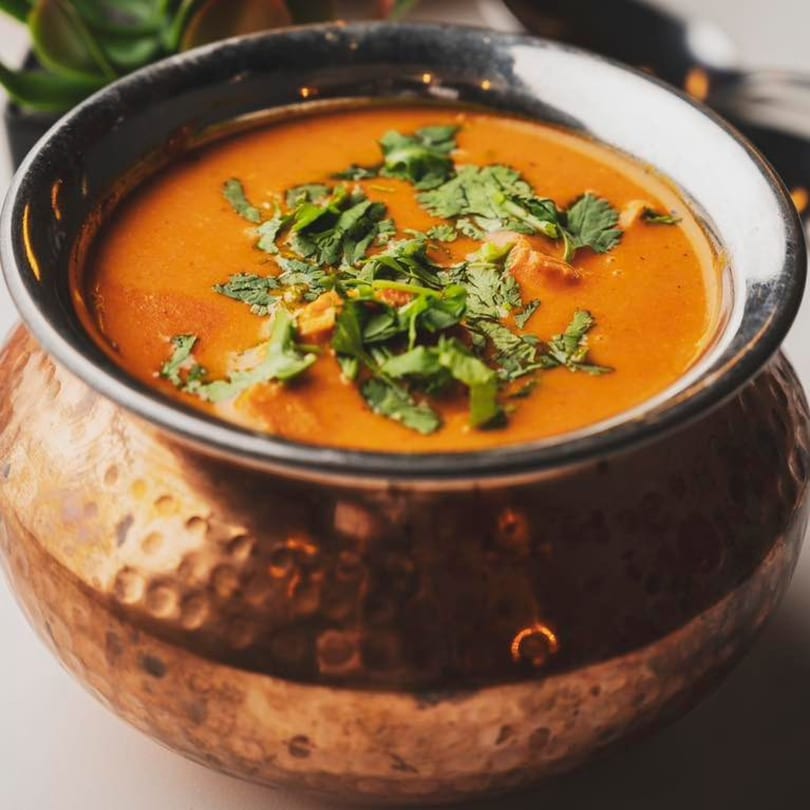 A copper bowl filled with vibrant orange Indian curry, garnished with fresh green cilantro.