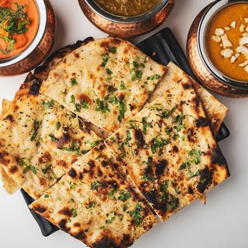 Garlic naan bread, cut into squares, served with three bowls of Indian curries.