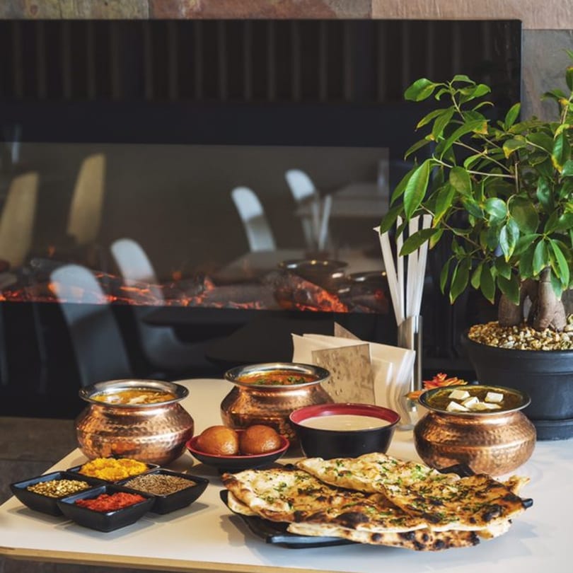 Indian dishes in copper pots, naan bread, spices, and dessert on a table by a fireplace.